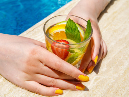 Woman hand holding glass with Citrus mint leaves infused water, cocktail, lemonade near pool. Summer iced cold drink with lemon, lime, orange, strawberry, mint on blue water brown marble backgroundの写真素材