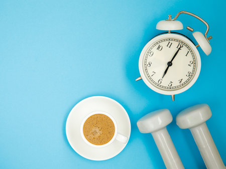 Top view on a pair of white dumbbell, cup of coffee and alarm clock on blue background. The concept of doing sports early in the morning. Time for self-training at home. Flat lay, copy spaceの写真素材
