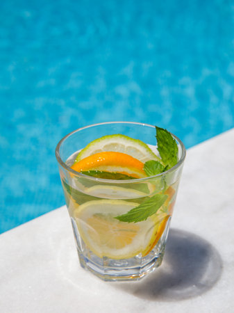 Citrus mint leaves infused water, cocktail, lemonade in glass near pool. Summer iced cold drink with lemon, lime, orange, mint on blue water white marble background, copy space, side view, verticalの写真素材