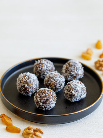 Healthy homemade energy balls: dry fruit ball cakes made from raisin, prune, dates, walnut, figs in black plate against white background, covered with coconut flakes. Close up copy space verticalの写真素材