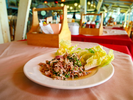 Thai food, spicy minced pork salad Larb Moo on white dish with spoon and fork on wooden background, with blurred thai cafe on the background, selective focusの写真素材