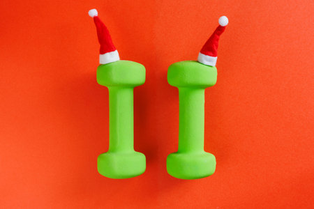 Composition with two Christmas green dumbbells in Santa hat on red background. Top view, flat lay, copy space close up minimalistic background, Gym Fitness New Year greeting cardの写真素材