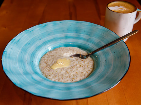 Healthy breakfast oatmeal porridge in blue bowl with spoon and cup of coffee on wooden table. Warm porridge oats with butter, vegan vegetarian weight loss dieting breakfast food. Close up.の写真素材