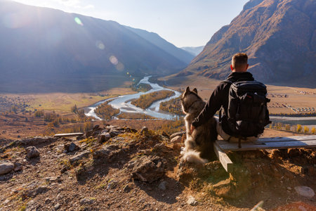 A man and his Alaska Malamut dog are taking a break from the hike. Sit down to enjoy the view of the Chulyshman river valley in Altai mountains in sunrise in autumnの写真素材
