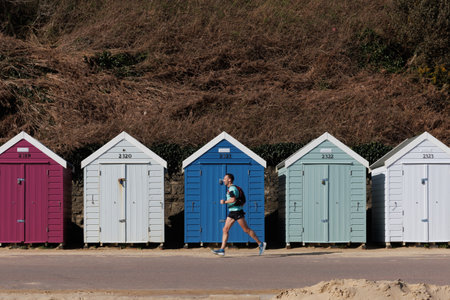 Active sport man running on seafront in beach with row of wooden beach huts on background in spring in Bournemouth, UK. South coast of England.の写真素材