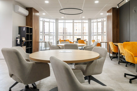 Empty working area in an open office. White round table surrounded by chairs in grey and yellow colors. Big window backgroundの写真素材