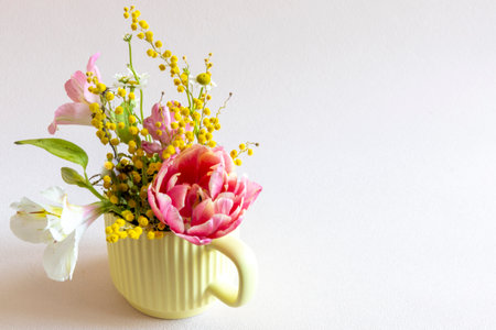 vibrant spring floral bouquet in soft yellow mug. pink tulip, white lily and yellow mimosa creating a cheerful atmosphere. Copy space, close up. Spring holiday, birthday, mother day greeting cardの写真素材