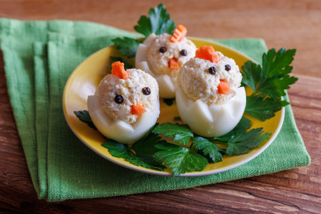 Fun and creative appetizer crafted three egg chick shape. Stuffed boiled eggs with tuna, carrot pieces, and peppercorns on yellow plate with parsley. Easter celebrationの写真素材