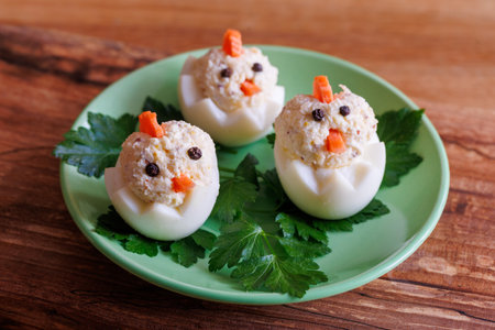 Fun and creative appetizer featuring three egg chick designs. Stuffed boiled eggs with tuna, carrot pieces, and peppercorns on green plate garnished with parsley. Easter foodの写真素材
