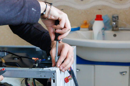 Hands using screwdriver on metal frame of washing machine, working on household appliance repair. Various cables and tools visible, emphasizing attention to detail and technical skillの写真素材