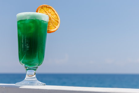 Refreshing green non-alcoholic cocktail with orange slice in glass with backdrop of blue sea. Perfect for summer relaxation and tropical getaways, travel holiday concept. Copy space.の写真素材