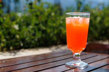 Bright orange drink served in glass with ice cubes on wooden table outdoor with blurred green foliage in background, summer concept, copy space.の写真素材