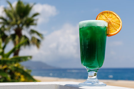 Vibrant green cocktail with an orange slice garnished on rim of a tall glass, set against tropical beach backdrop with palm trees and clear blue ocean. Copy space. Holiday travel.の写真素材