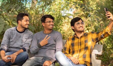 Group of friends taking a self portrait in the park - Concept of young people addicted to selfie - Three friends are sitting on bench in park and taking selfie at Park.の写真素材