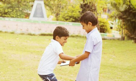 Two kids fighting for tablet at park outdoor - Liite boy trying to grab a tablet from her friend or brother - Concept of children's addiction, aggression on electronic devices or gadgetsの写真素材
