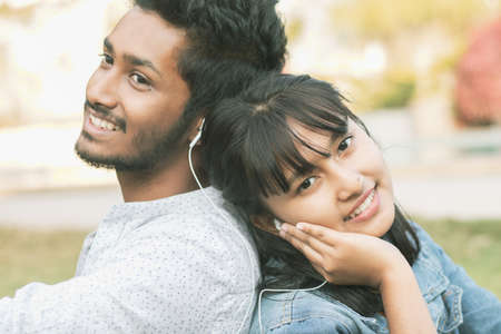 Young teenage friends enjoying listening to favorite song or music by Sitting back to back - Concept of couple relaxing, picnic in park, Love, dating, romance and modern lifestyle.の写真素材