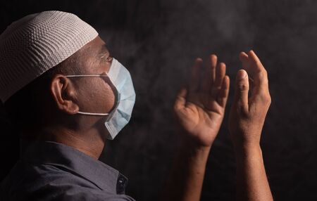 Muslim Man with medical mask and cap praying to god in dark room to protect or save from covid-19 or...