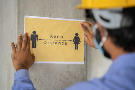 worker pasting Keep 6 feet distance on wall at work place or construction site due to coronavirus or covid-19 pandemic - concept of safety measures, back to work, new normal and protection.の写真素材