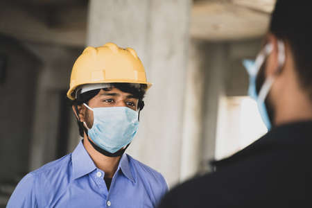 Two Construction workers or engineers at site talking by wearing medical face mask while maintaining...