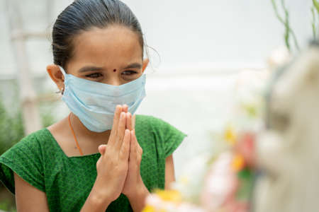 Young girl kid in medical mask praying to god to protect from coronavirus or covid-19 by folding hands in namaste gesture.の写真素材