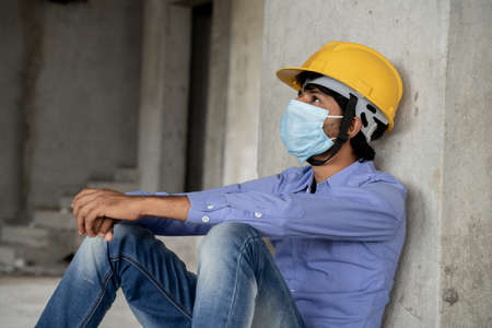 Head shot side view of construction worker seeing outside from construction building window in sad - Industrial worker in a hardhat with medical mask due to coronavirus or covid-19 crisis.の写真素材