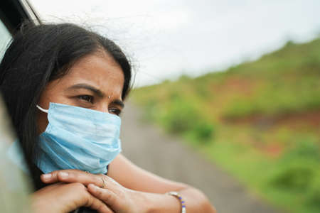 Sad Young girl in medical mask watching out from car window - concept of travel, vacation and road...