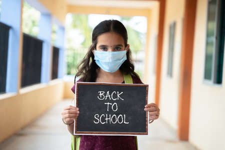 Young girl in medical mask holding back to school signage board at corridor - concept of school...