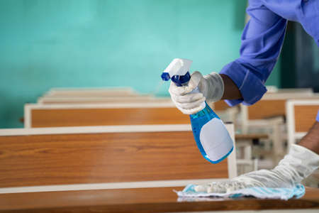 Close up of hands with gloves disinfecting desk by using sanitizer at classroom - Cleaning dust on table surface with cloth and Disinfectant Spray to protect from coronavirus or covid-19 infection.の写真素材
