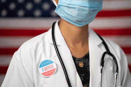 Doctor or nurse medical mask and stethoscope wearing placing I voted sticker to her coat - concept of US election with US flag as backgroundの写真素材