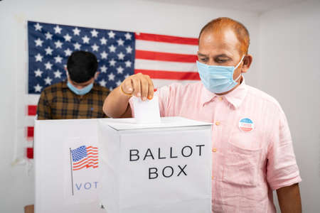 Man in medical mask placing ballot paper inside the ballot box - Concept of in person voting and...