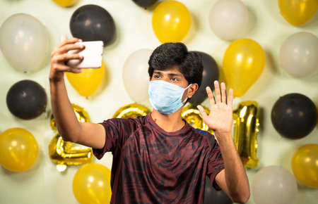 Young man with medical mask busy making video call on mobile over 2021 new year decorated background...
