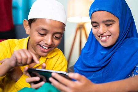 Two muslim sibling kids busy using mobile at home - concept of kids using technology, internet and...