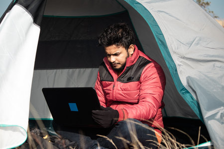 Young man busy working on laptop inside the tent during camping - concept of digital nomadic lifestyle and solo travel.の写真素材