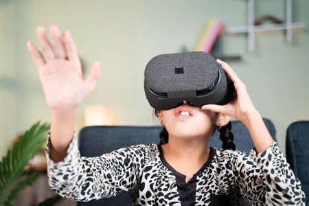 Young girl kid with VR or virtual reality goggles feeling or enjoying the 360 degree virtual environment at home - Concept of showing modren VR technology in modern lifestyleの写真素材