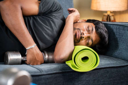 Tired young man sleeping on sofa by holding dumbbells and yoga mat - concept of healthcare,...