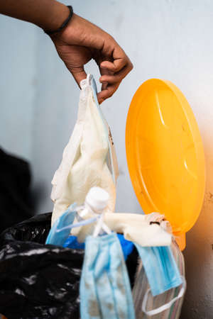 Close up of healthcare worker or doctor hands throwing single use disposable medical face mask and gloves into filled trash can or garbage bin at hospital during coronavirus covid-19 pandemic.の写真素材