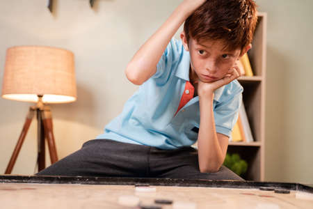Overthinking Confused Caucasian kid scratching head to decide which coin to hit while playing carrom...