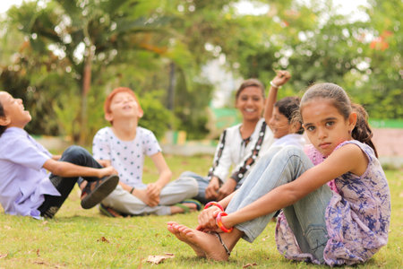 Group of kids bulling girl at park during summer camp - Little girl looking camera, sitting lonely while friends playing and having funの写真素材