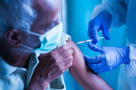 Senior elderly patient with mask getting vaccination or jab at hospital - Doctor in protective Hazmat suit treating Covid patient injecting anti-viral vials at covid care Centreの写真素材