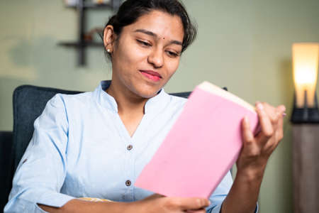Happy young businesswoman reading book or studying during leisure time or for examination at home by sitting on sofa at home - concept of reading hobby.の写真素材