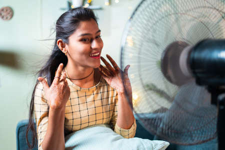 Happy smiling Indian Woman enjoying fan air during to heat stroke while sitting on couch - concept of heat wave or feeling cool air from fan at homeの写真素材