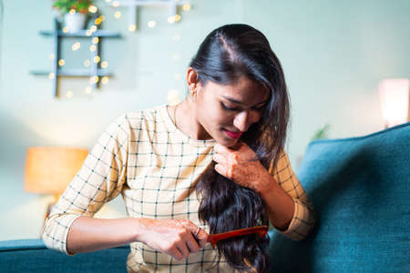 Young Indian girl on sofa combing her long hairs - concept of woman getting ready and daily ritualsの写真素材