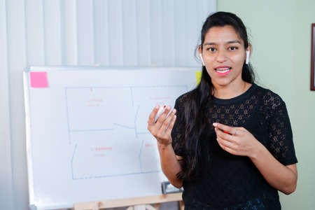 young girl busy explaining from board during online class by looking camera at home with wireless headphones - Concept of e-teaching,Remote learning,virtual education during coronavirus outbreakの写真素材