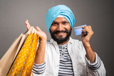 Happy Young Sikh man with shopping bag and credit card in hand looking into camera on studio background - concpet of online e-commerce festival shoppingの写真素材