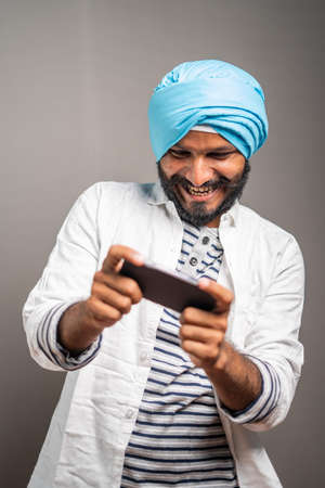 Young smiling sikh man busy playing video game on mobile phone - concept gaming addiction, using modern technology on studio backgroundの写真素材