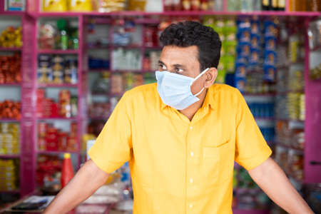Merchant with mdical face mask at groceries or Kirana store waiting for customers by looking around during coronavirus covid-19 pandemic - concept of small businesses reopen and new normal lifestyle.の写真素材