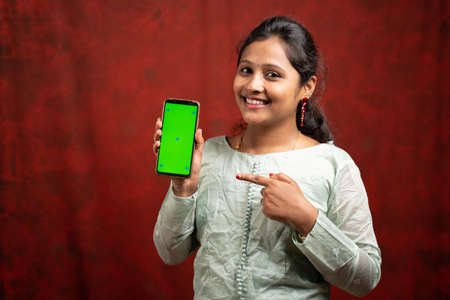 Happy smiling Indian girl in ethnic dress Pointing finer to mobile phone with mock up green screen for Diwali festival advertisement by looking at cameraの写真素材