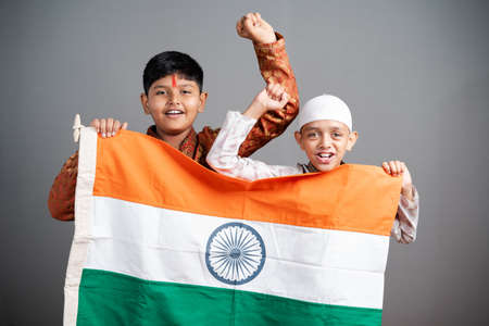 Happy Hindu Muslim kids shouting as India by holding Indian flag on gray background - concept of...