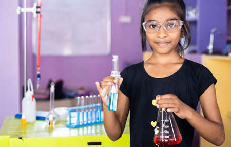 Happy confident school kid at science lab with chemical flask in hand looking camera at classroom - concept of creativity, child prodigy and educationの写真素材