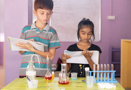 Two diverse kids busy making science chemistry laboratory experiment by referring manual - concept showing of education, friendship, socializing at schoolの写真素材